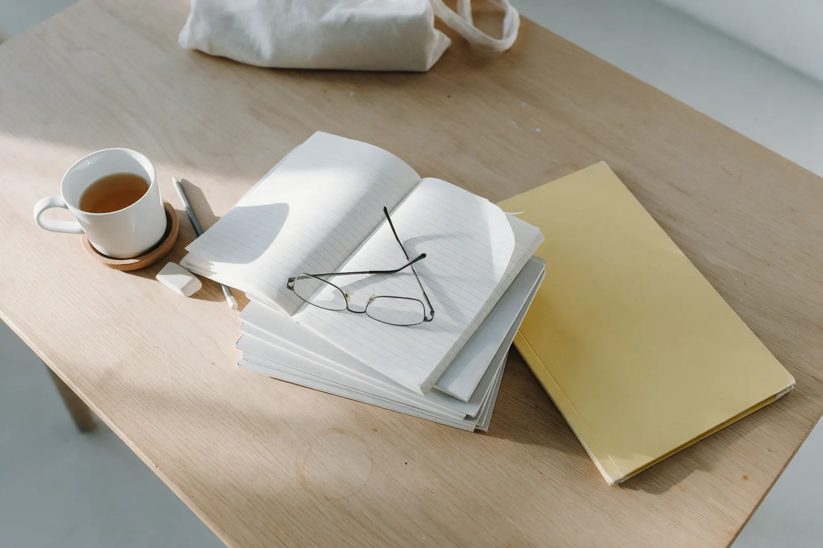 A light oak desk seen from above with a stack of exercise books, the top one open on blank ruled pages with reading glasses on it, a yellow card folder, a mug of tea on a cork coaster, a pencil and an eraser