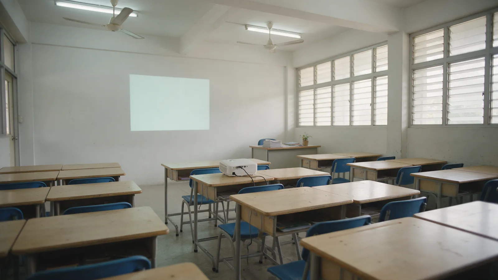 An empty classroom seen from the back row, desks with blue chairs, a pale blank projected rectangle on the front wall