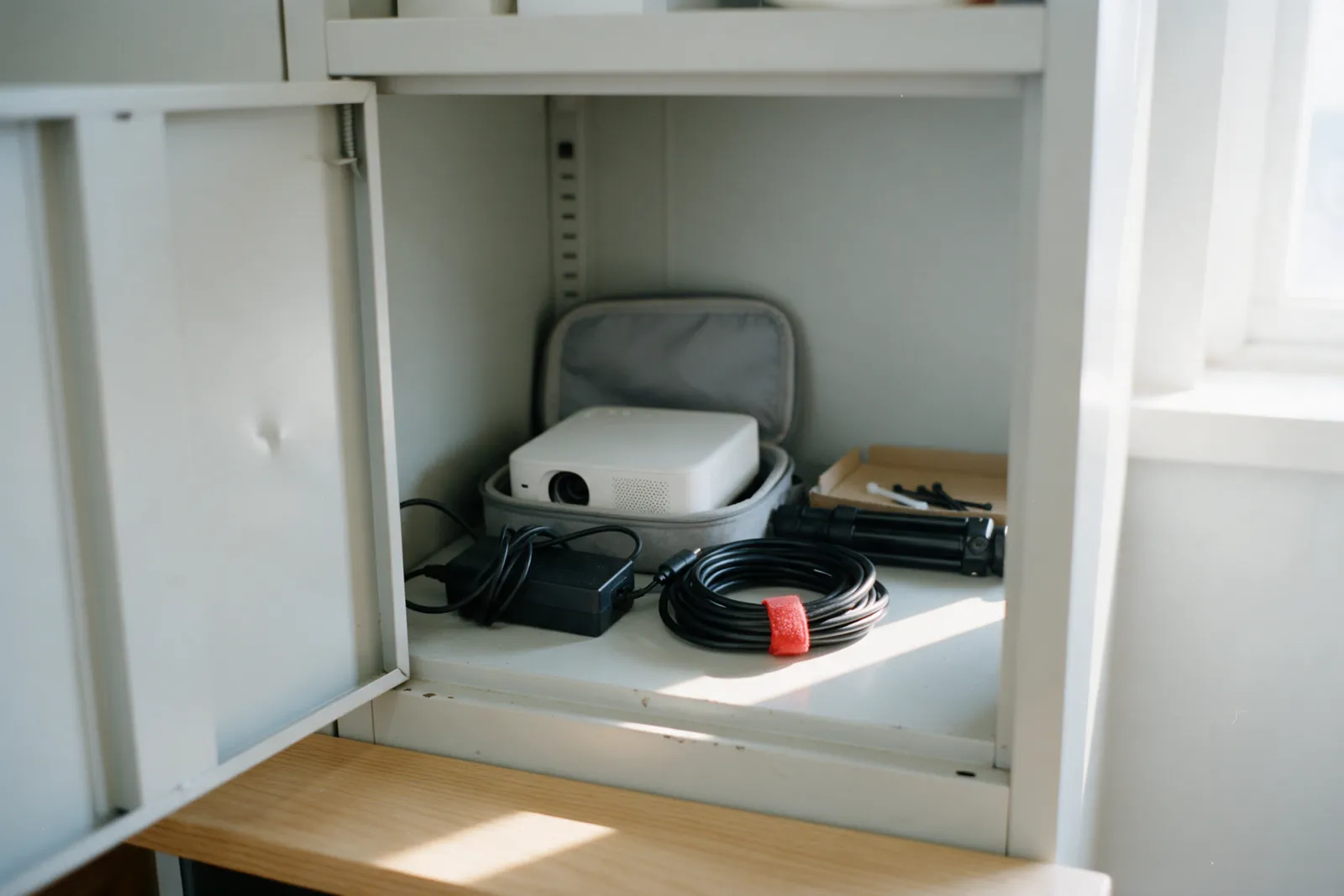 A small white portable projector in an open grey case on a cupboard shelf, with its power adapter, a coiled cable and a folded tripod