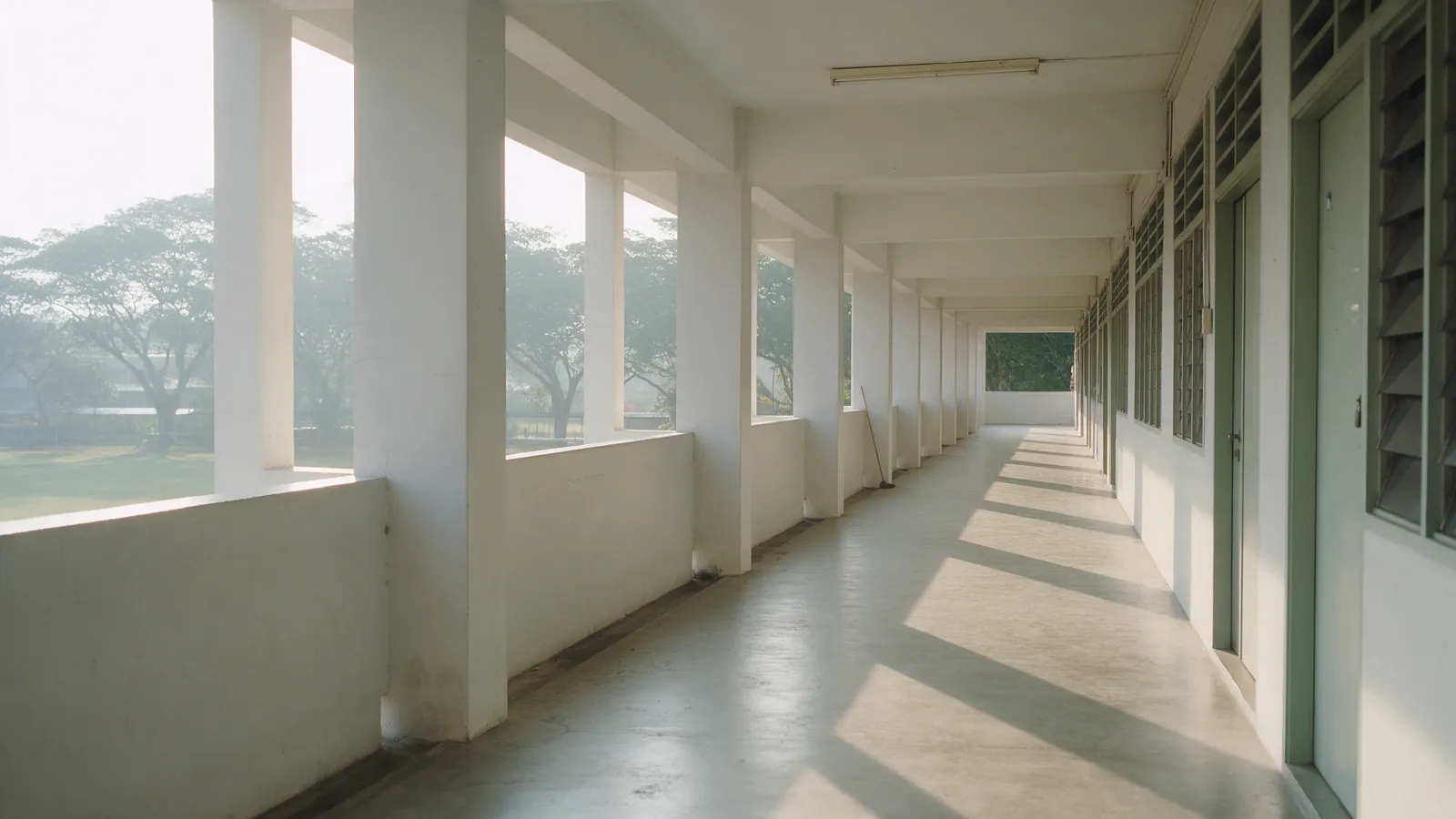 An open-air school corridor with white columns along one side and classroom doors along the other, morning light across the floor